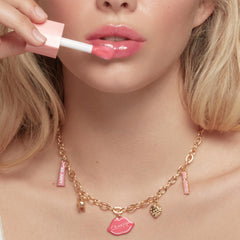Brooklyn Charm x ColourPop Necklace showcasing a gold link chain with pink enamel charms - lip-shaped ColourPop pendant, mini gloss tube, heart and sparkly charms - worn by a model applying pink lip gloss in a studio close-up; ColourPop Necklace.
