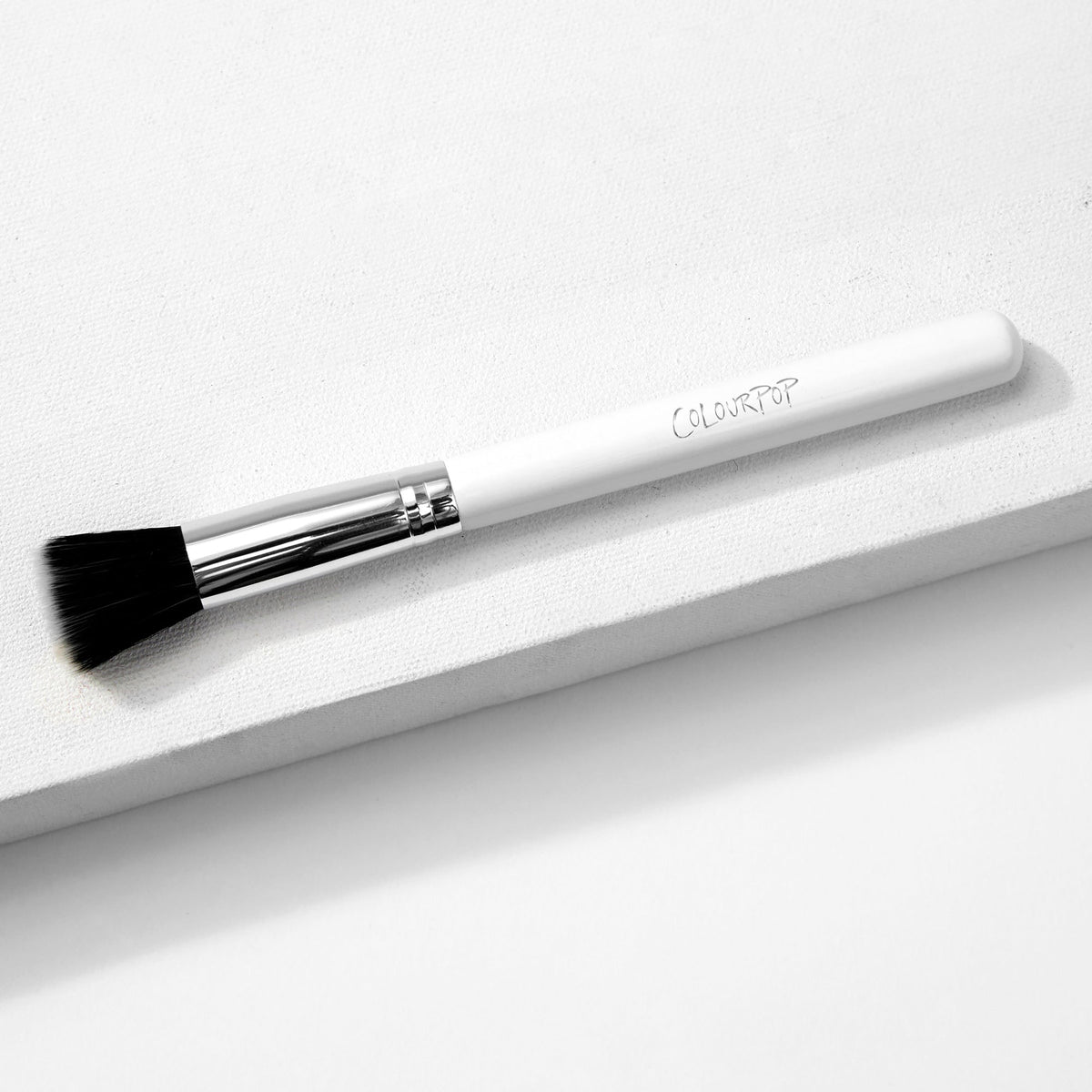 ColourPop stippling makeup brush resting diagonally on a white textured block, featuring duo-fiber black-and-white bristles, a shiny silver ferrule, and a white handle with the ColourPop logo.