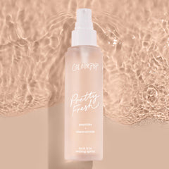 ColourPop Pretty Fresh Setting Spray, Lock It In, stands upright with a white pump on a frosted blush bottle labeled peptides + niacinamide, casting a soft shadow against rippled beige water for a clean, dewy makeup-locking look.