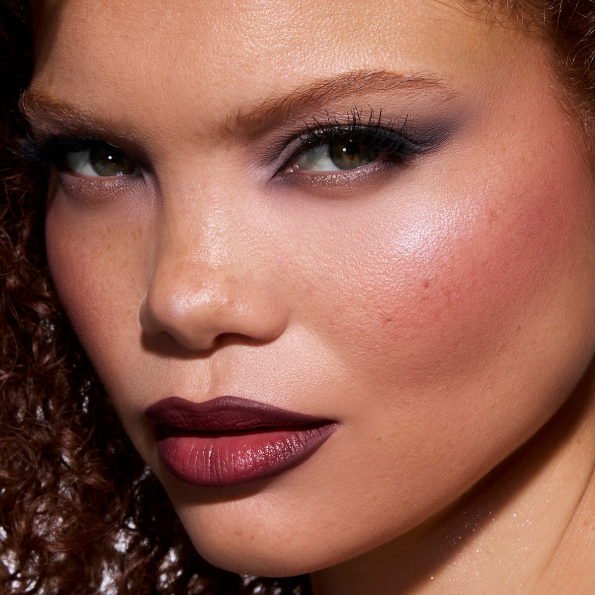 Model wearing Change Me Mega Highlighter from the Twilight collection, showcasing a luminous pink-lilac glow on the cheekbone with smoky eye makeup and deep berry lipstick in a close-up beauty shot.