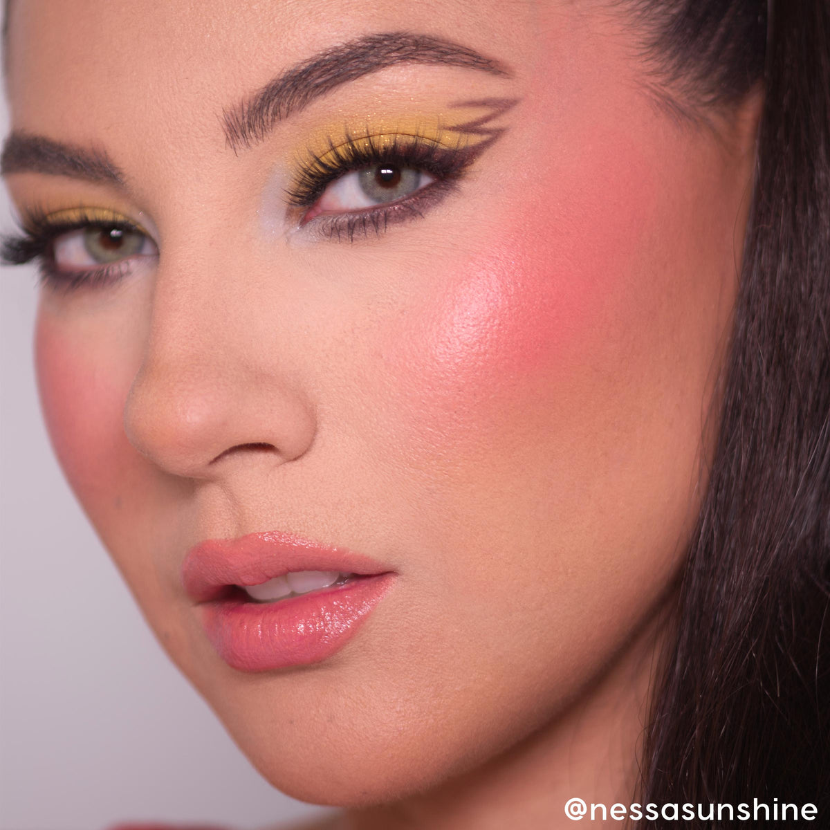 Electrifying Dreams ColourPop Cream Blush delivering a dewy coral-pink glow on high cheekbones, shown in a close-up face with bold yellow graphic winged eyeliner, long lashes, and glossy pink lips under soft studio lighting.