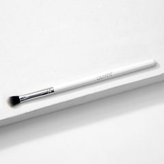 ColourPop Blending Brush makeup brush resting diagonally on a white textured surface, featuring fluffy angled black-to-white bristles, a silver ferrule, and a slim white handle, made for seamless eyeshadow blending.